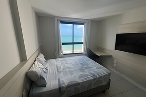Un dormitorio con cama, televisión, ventana y vista al mar, además de una silla.
