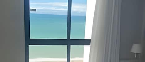 A hotel room with a bed, a window with a view of the sea, and a nightstand with a lamp.
