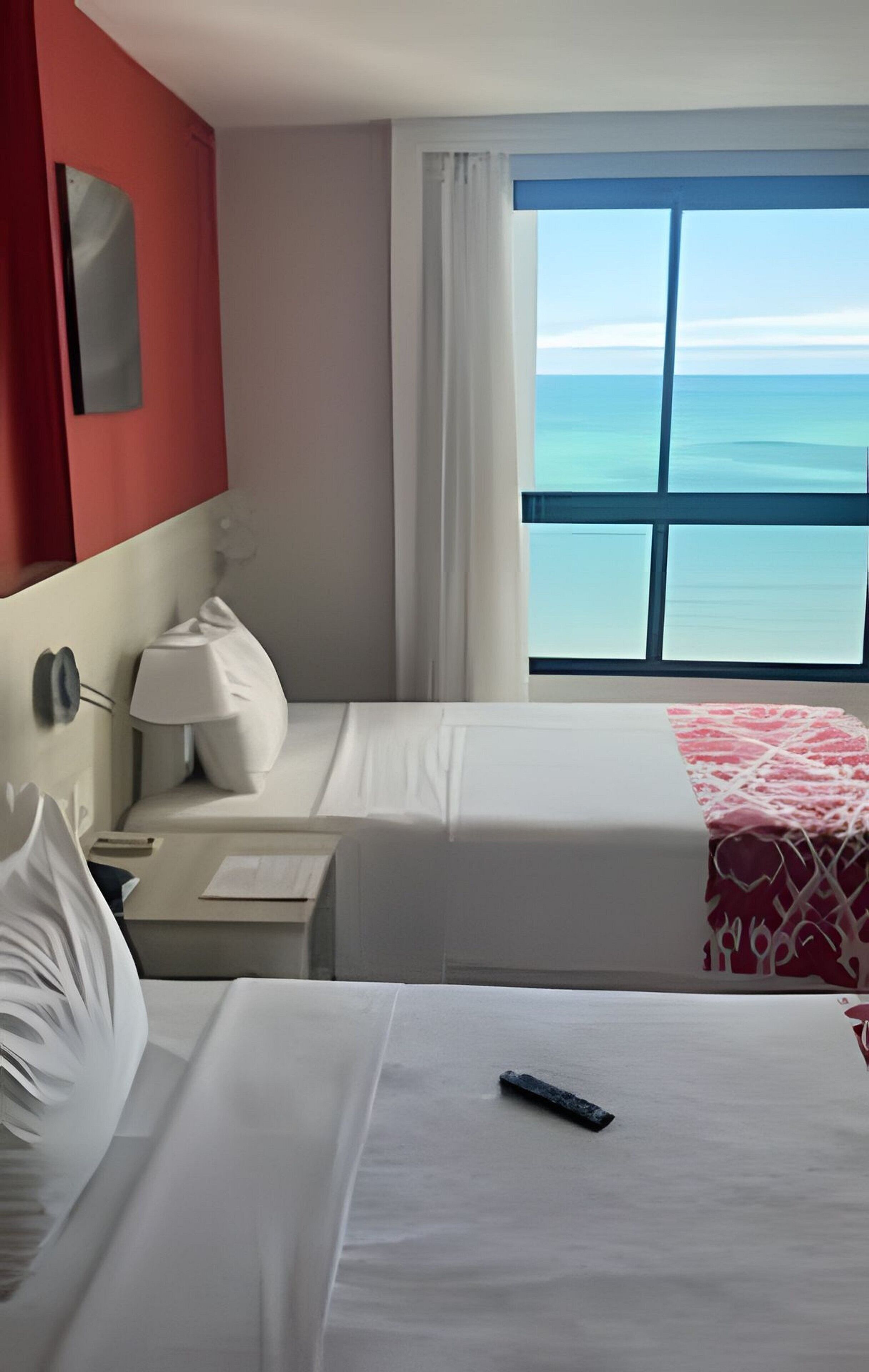 A hotel room with two beds, a nightstand, a lamp, a window with a view of the ocean, and a red accent wall.