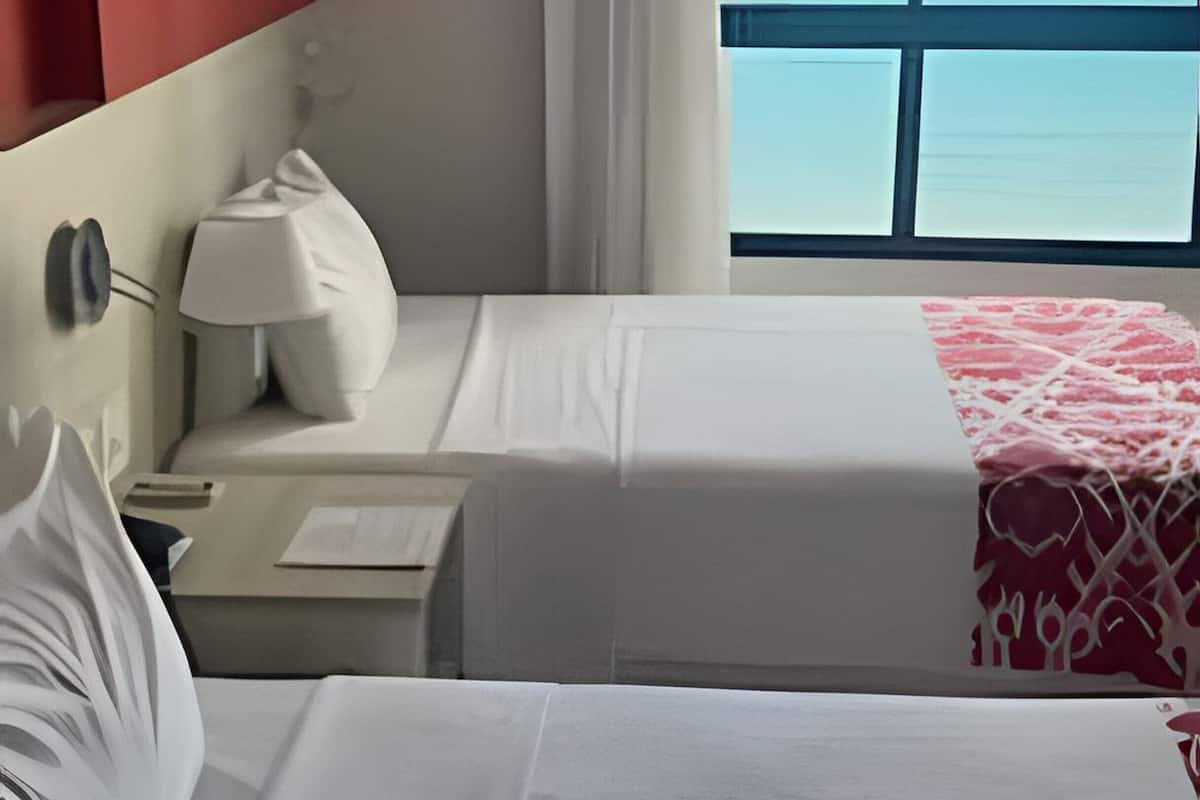 Hotelkamer met twee bedden, een nachtkastje, een lamp, een raam met uitzicht op de oceaan, en een rode accentmuur.