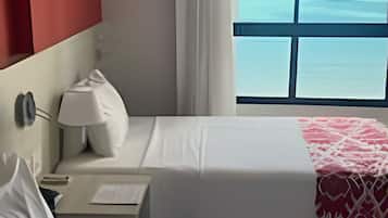 A hotel room with two beds, a nightstand, a lamp, a window with a view of the ocean, and a red accent wall.