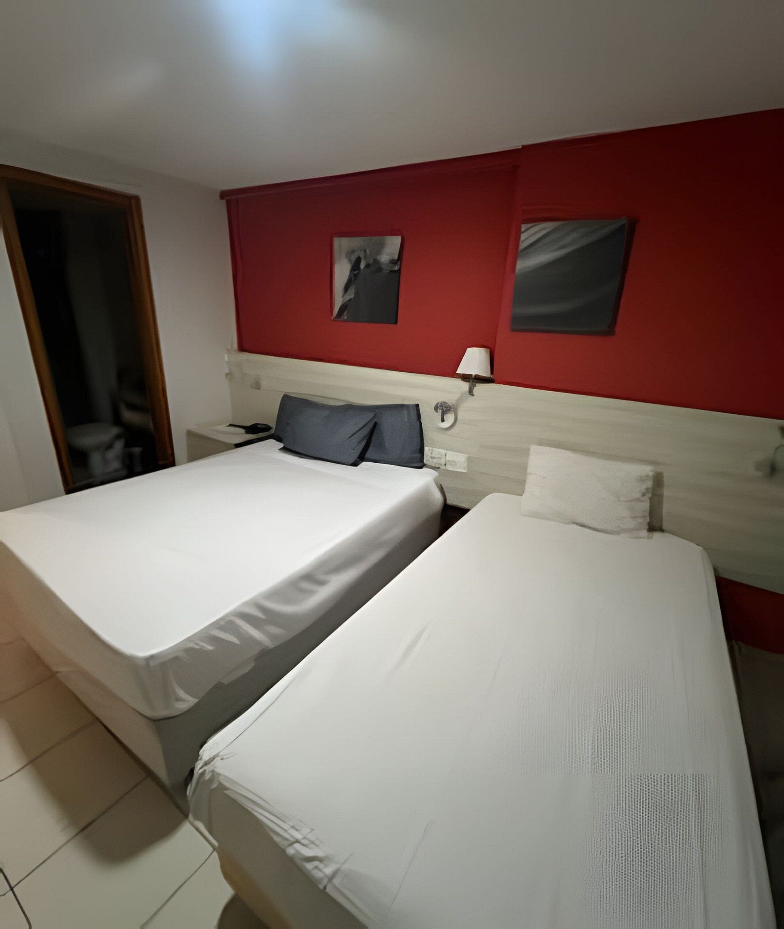 A hotel room with two beds, a red accent wall, a nightstand with a lamp, and a door leading to a bathroom.