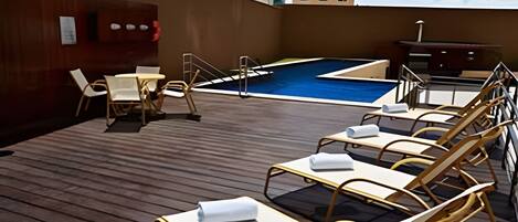 Et tagområde med swimmingpool, liggestole og et lille bord, omgivet af en træterrasse.