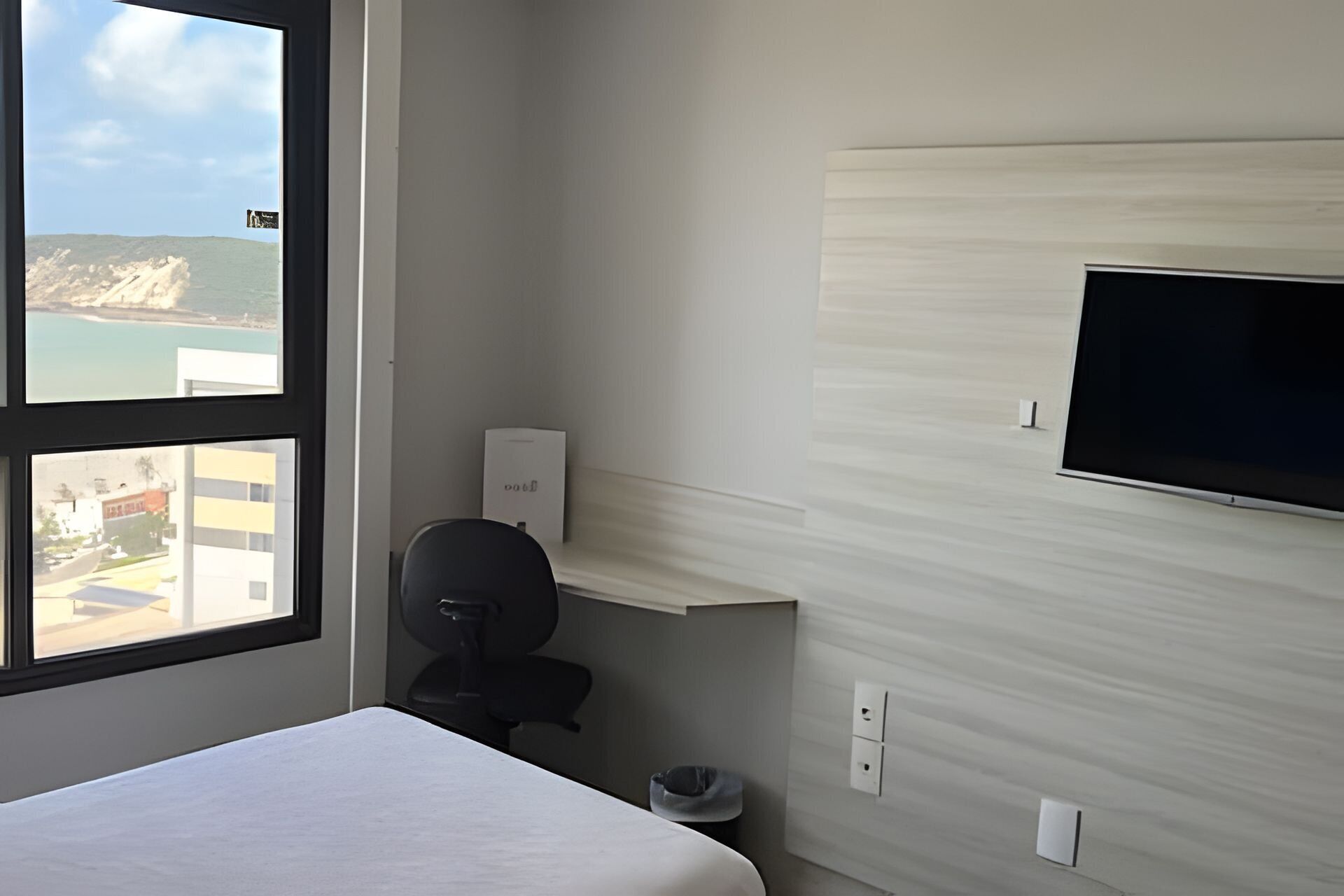 Une chambre d’hôtel équipée d’un lit, d’un bureau, d’une chaise, d’un téléviseur fixé au mur et d’une fenêtre donnant sur une plage et des immeubles.
