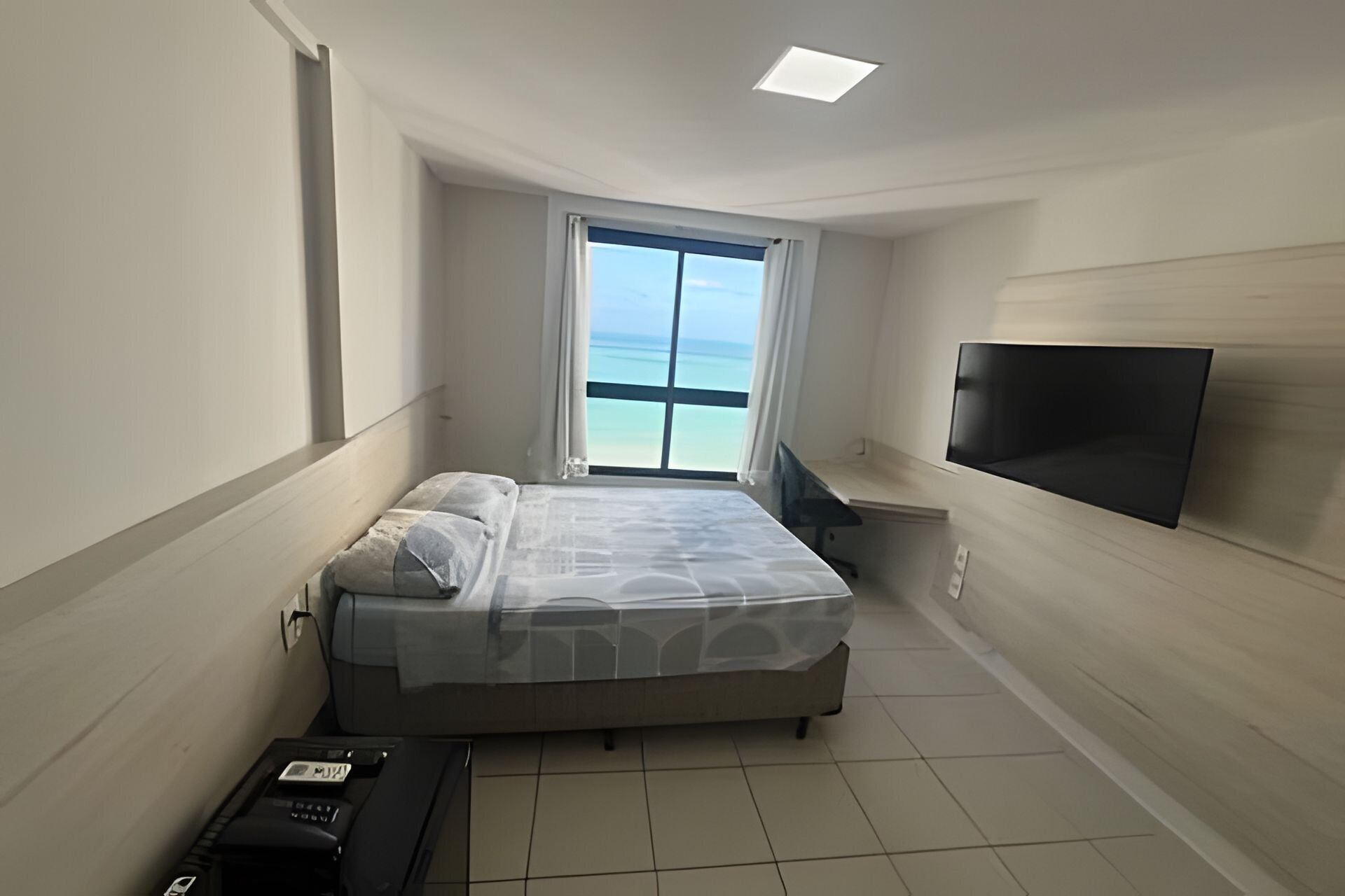 A bedroom with a bed, a TV mounted on the wall, a window with a view of the sea, and a desk with a chair.