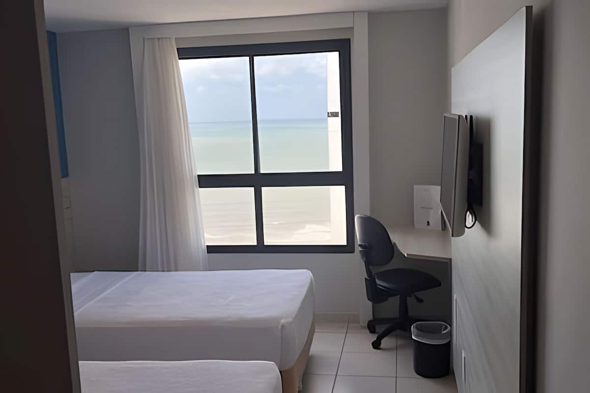 Een hotelkamer met twee bedden, een bureau, een stoel, een televisie en een groot raam met uitzicht op het strand.