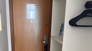 Un couloir avec une porte en bois, une petite étagère sur laquelle se trouve un objet noir, et un mur blanc.