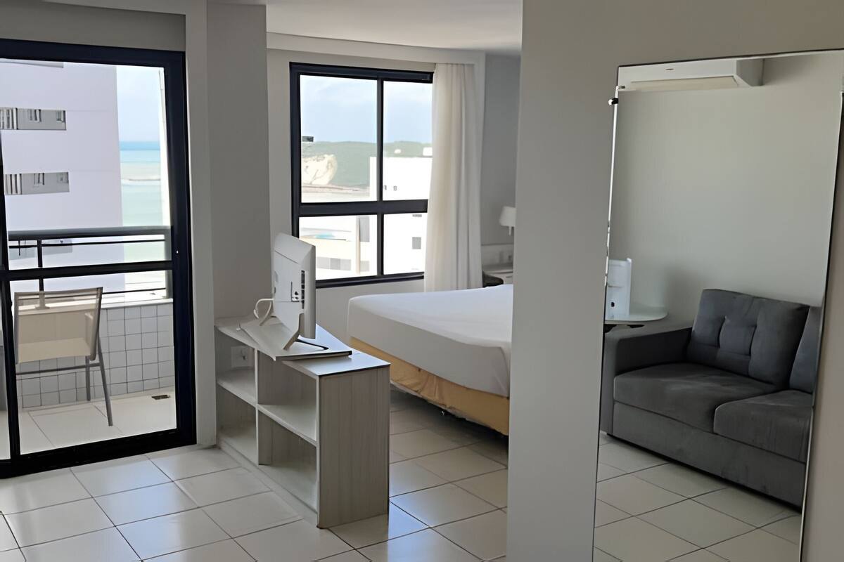 A hotel room with a bed, a desk with a computer, a sofa, a balcony with a view of the sea, and a window with curtains.