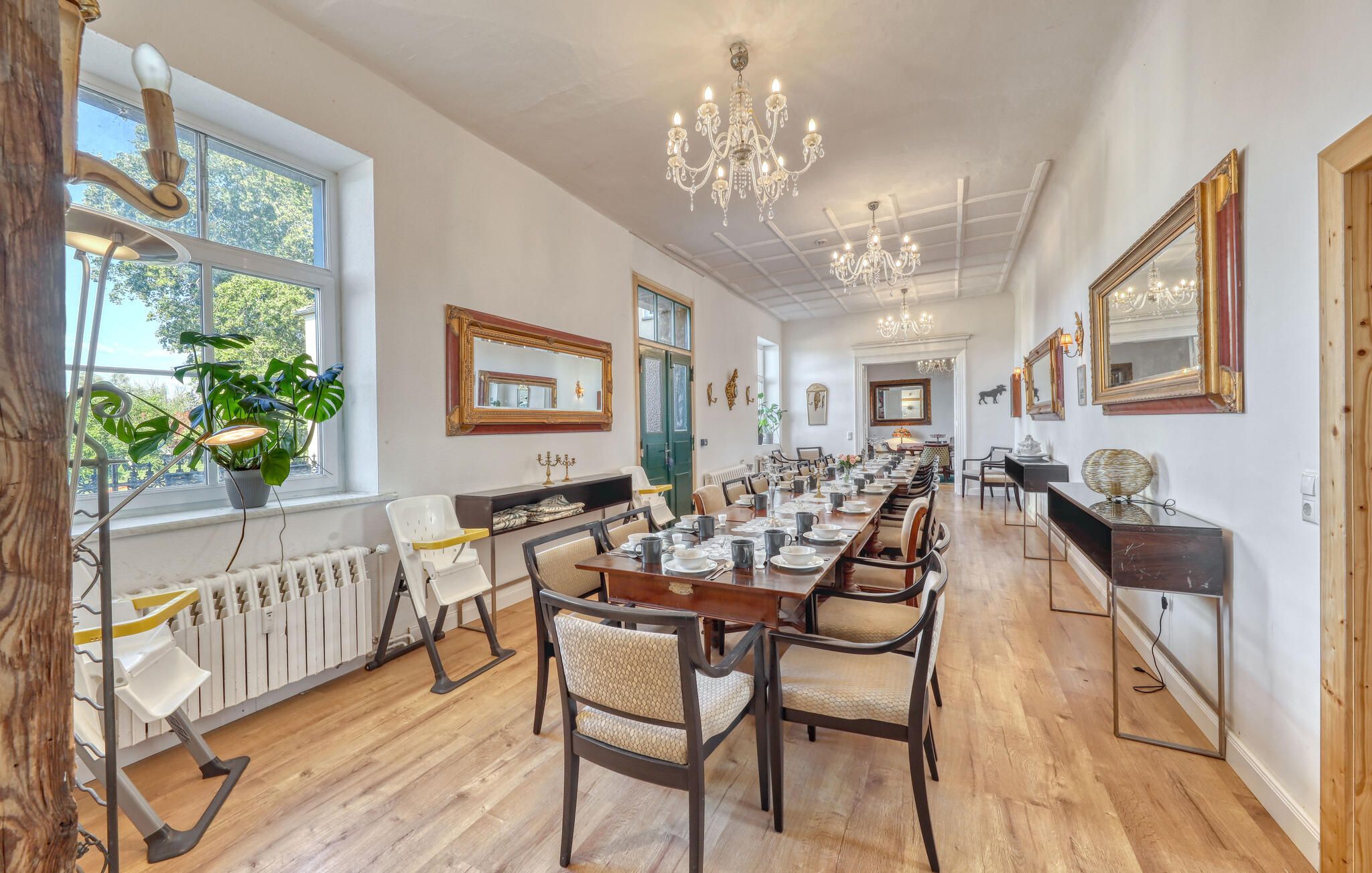 A long dining table with chairs, chandeliers, and large windows with plants.