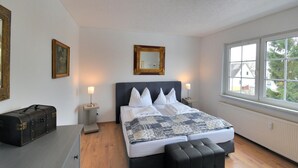 Quarto com uma cama grande, duas luminárias de cabeceira, cômoda, baú e janela com vista para árvores e edifícios.