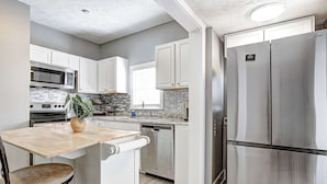 A modern kitchen with white cabinets, stainless steel appliances, and a wooden dining table. - The Yellow Door – Cozy Indianapolis Retreat (Indianapolis)