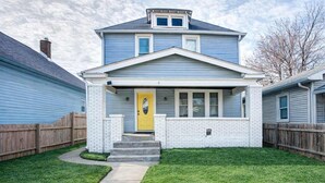 A two-story house with a front porch, a yellow door, and a brick foundation. - The Yellow Door – Cozy Indianapolis Retreat (Indianapolis)