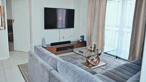 A modern living room with a large flat-screen TV mounted on the wall, a glass coffee table, and a grey sofa.