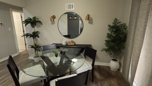 Un comedor con mesa de vidrio, sillas negras, un espejo redondo y dos plantas en macetas.