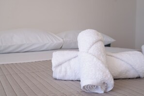 Ein ordentlich gemachtes Bett mit weißen Leinen und Kissen, ein aufgerolltes Handtuch und ein aufgeräumtes, minimalistisches Zimmerdesign.