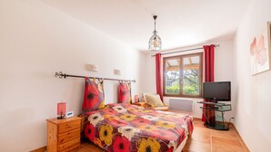 A bedroom with a floral bedspread, a wooden nightstand, a TV on a stand, and a window with red curtains. - Villa La Bulle - Piscine Privée (Grambois)