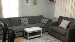 Een woonkamer met een grijze bank, een klein bijzettafeltje en een kamerbreed tapijt. Er staan twee stoelen, een plafondventilator en er hangen kunstwerken aan de muren.