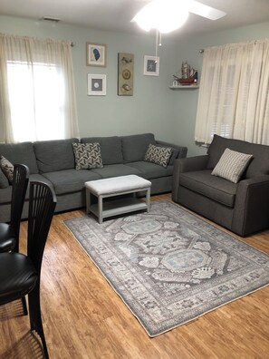 A living room with a grey sofa, a small coffee table, and a patterned rug. There are two chairs, a ceiling fan, and framed artwork on the walls. - Perfect 2-bedroom plus sleeping porch house with AC in peaceful sleeps 6 (Lavallette)