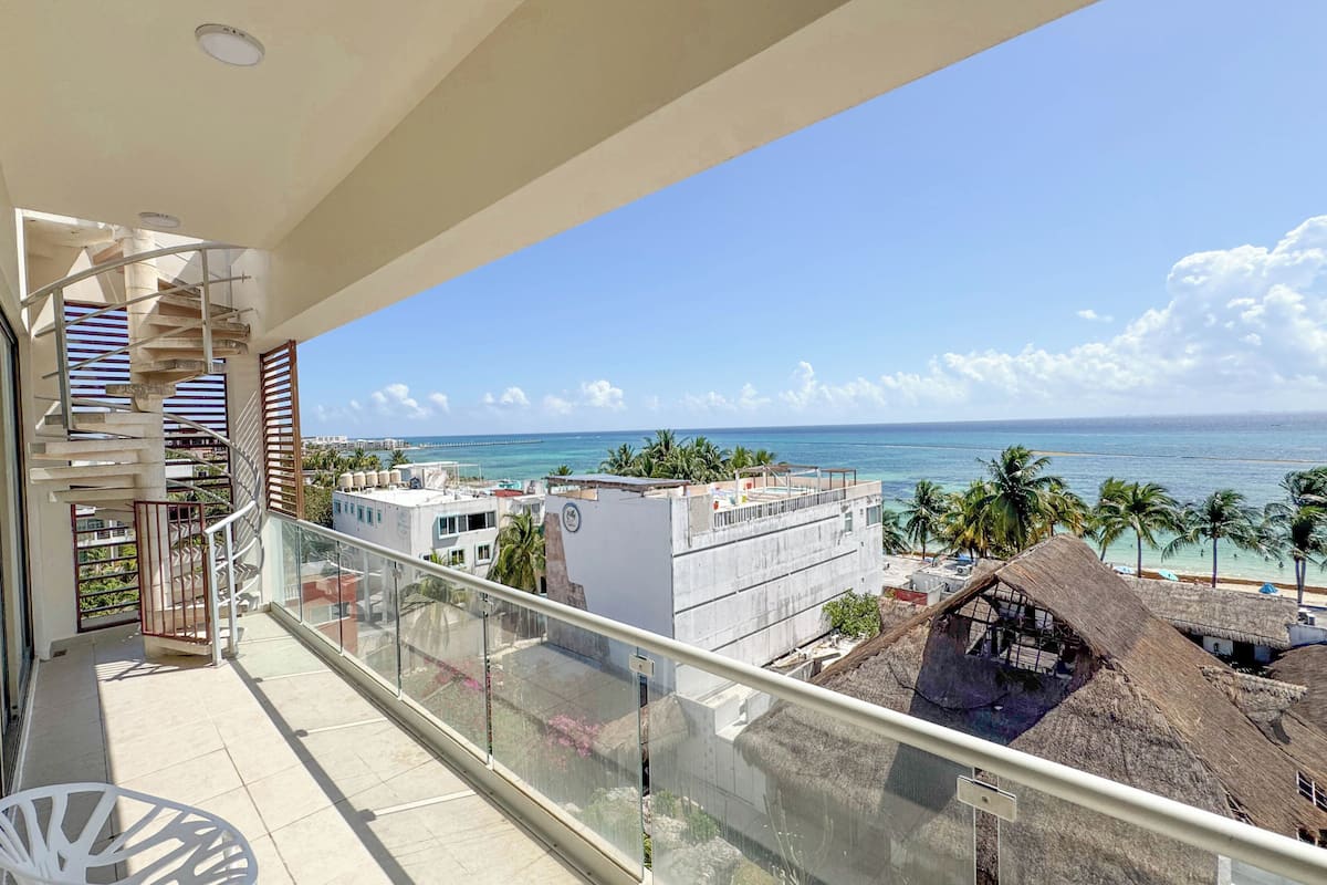 A balcony with a view of the sea, a thatched-roof structure, and a building with water tanks.
