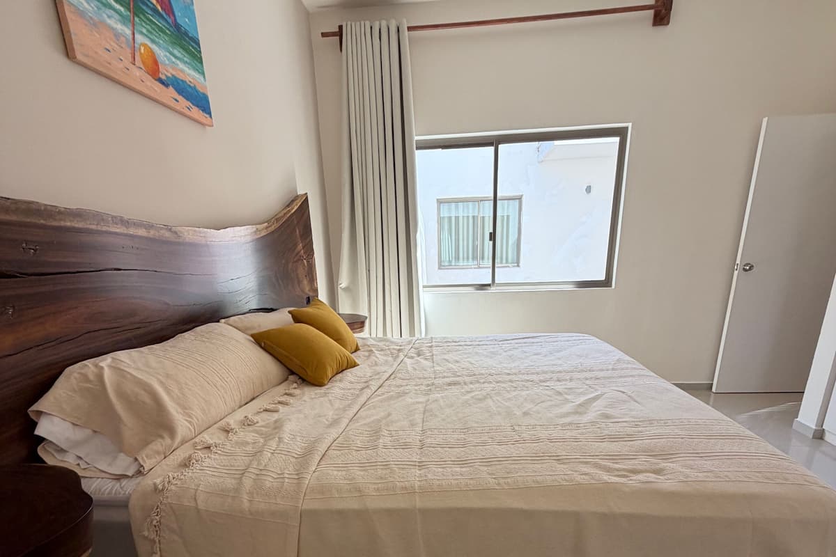 A bedroom with a wooden headboard, a large bed with a beige bedspread, two mustard pillows, a window with curtains, and a door.