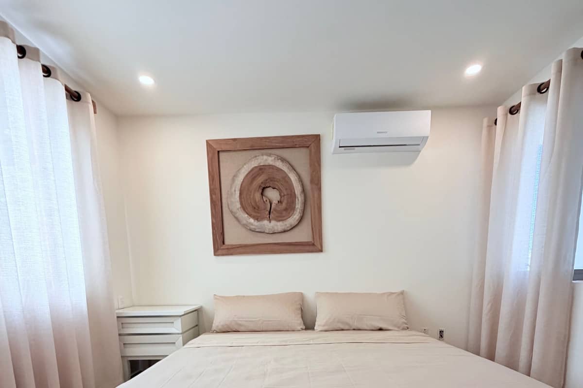 A bedroom with a large bed, a bedside table, a wall-mounted air conditioner, and a framed picture.