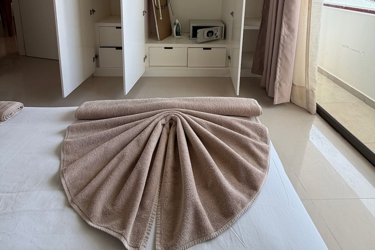 A bedroom with a bed, a wardrobe with open doors, a window with curtains, and a view of buildings outside.