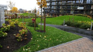 Een aangelegde tuin met een verhard pad, een houten speelconstructie en een modern appartementencomplex op de achtergrond.