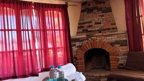 Ein Zimmer mit Kamin aus Stein, roten Vorhängen, einem Bett mit rot-weiß gestreifter Bettdecke und einem grünen Korb mit Wasserflaschen und einer Papiertuchrolle.