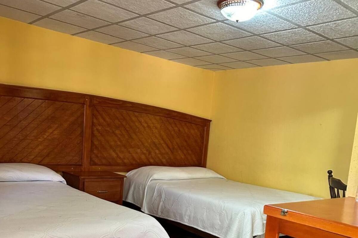 A room with two beds, a wooden headboard, a small table, and a ceiling light fixture.