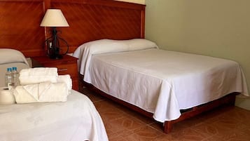 Habitación de hotel con dos camas, cabecero de madera, mesita de noche con lámpara y zona de baño.