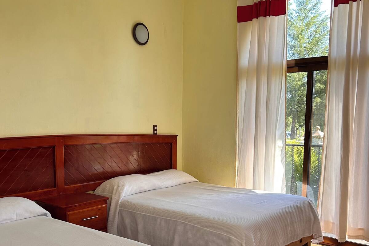 A room with two beds, wooden headboards, a bedside table, a clock on the wall, and a window with curtains.
