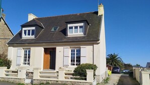 Una casa de dos pisos con techo oscuro, contraventanas blancas y puerta de madera.