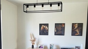 Un comedor con una mesa rectangular, dos sillas negras y una silla blanca. Sobre la mesa, una lámpara colgante negra con cuatro bombillas. Las paredes están decoradas con obras de arte enmarcadas.