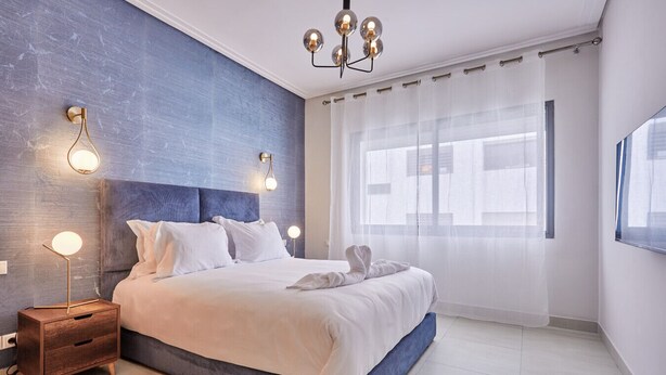 Une chambre à coucher moderne avec un grand lit, une table de chevet en bois, un luminaire fixé au mur et des rideaux légers.