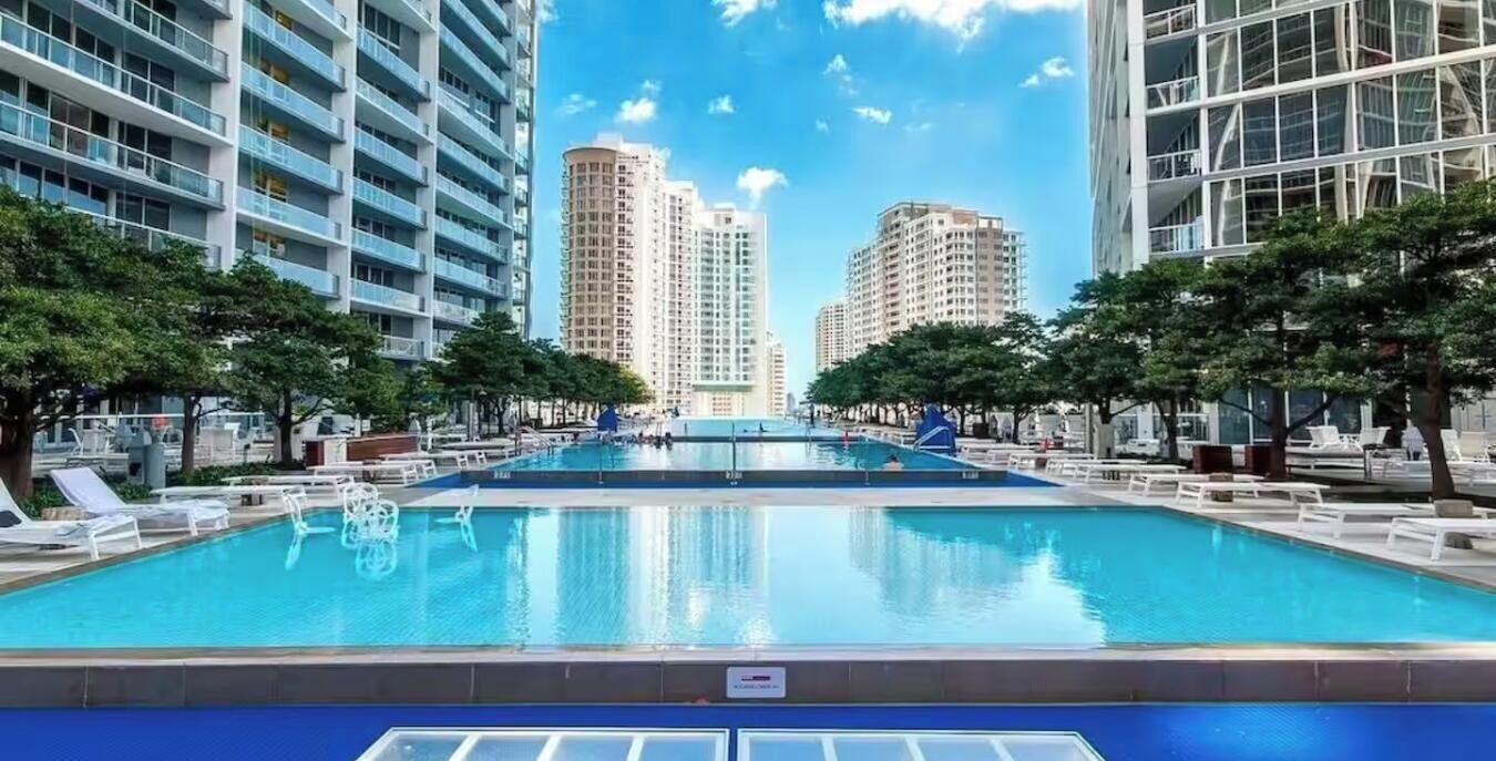 A swimming pool surrounded by modern buildings, trees, and lounge chairs.