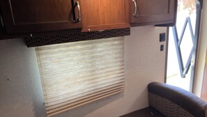A compact RV interior with wooden cabinets, a window with blinds, a cushioned bench, and a visible door. - Cozy RV Camper in tranquil Brandon near everything (Brandon)