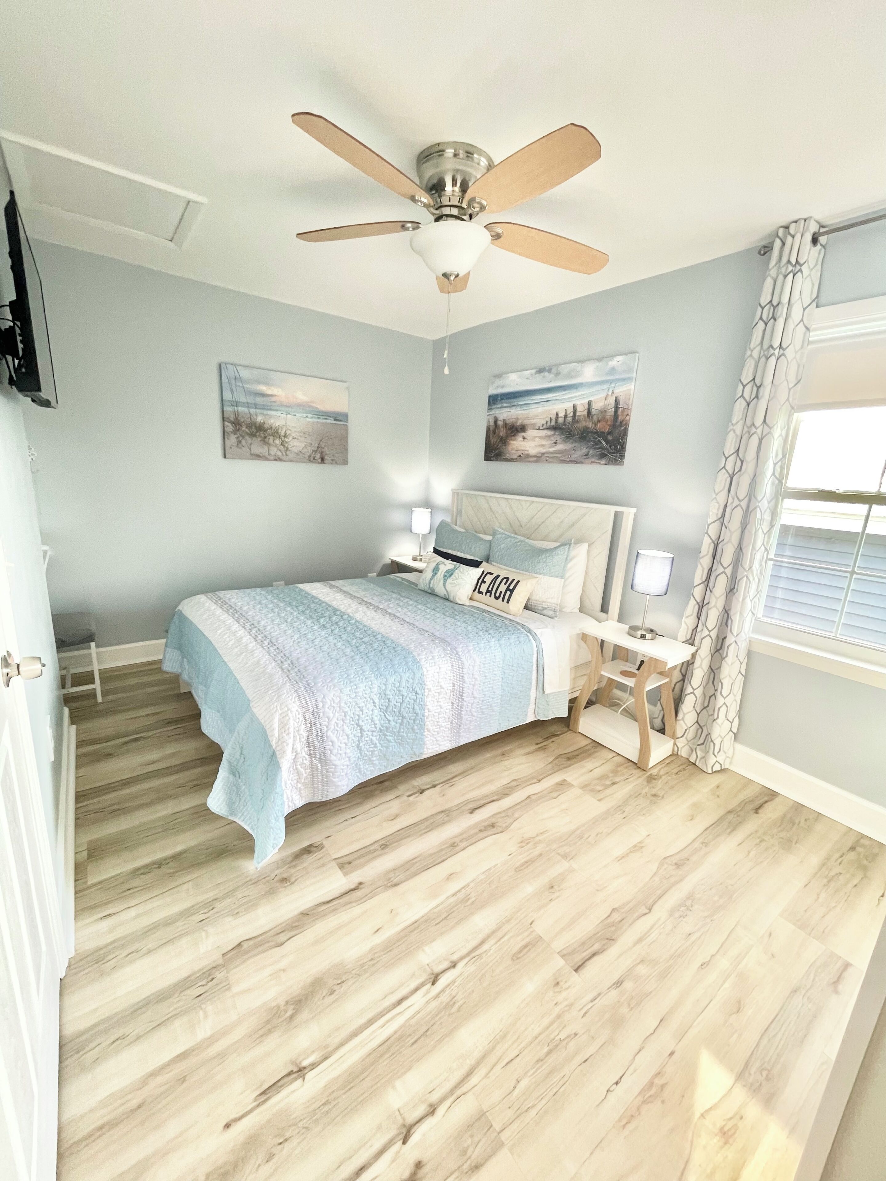 A bedroom with a bed, a ceiling fan, a nightstand, a lamp, and two wall-mounted paintings.