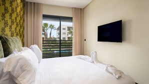 Ein Hotelzimmer mit einem großen Bett, weißer Bettwäsche, einem Flachbildfernseher an der Wand und Blick auf Palmen und Gebäude durch das Fenster.
