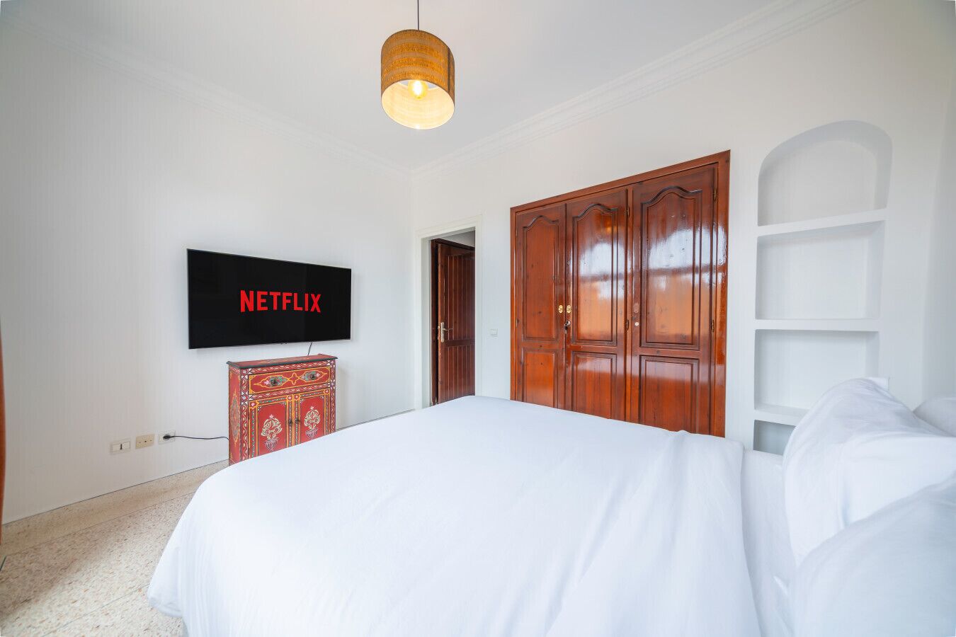 Une chambre à coucher avec un grand lit, une armoire, un téléviseur fixé au mur et un luminaire suspendu.