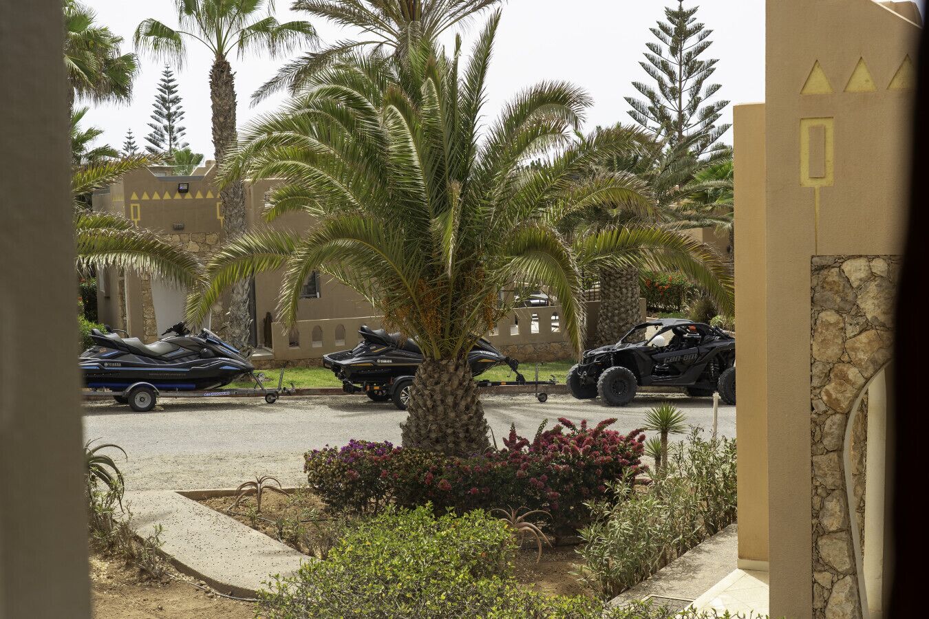 A parking area with several vehicles, including a jet ski, a motorcycle, and an ATV, surrounded by palm trees and a stone wall.