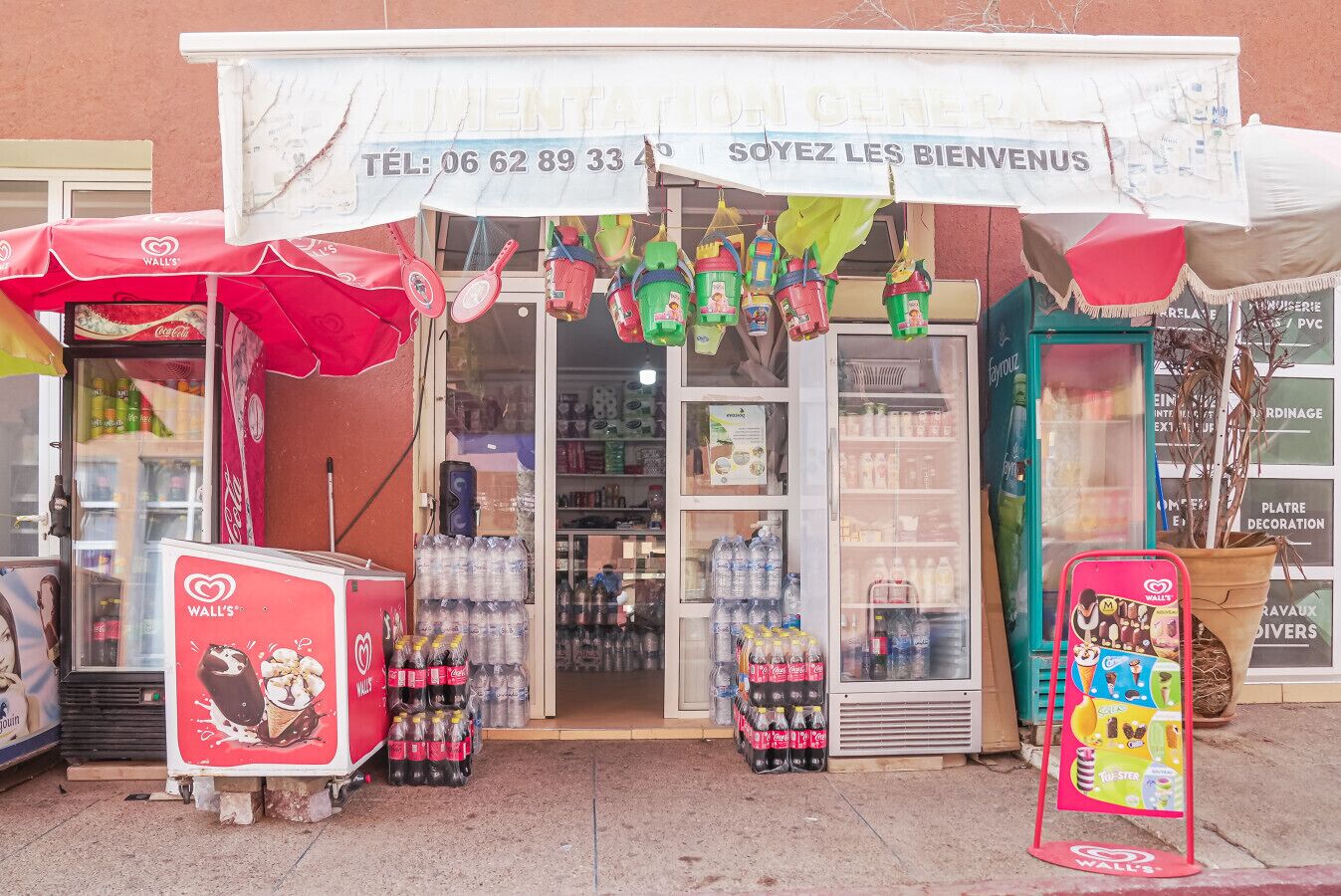 A storefront with a sign reading "SOYEZ LES BIENVENUS" and a phone number. There are vending machines outside, and a display of bottled water and snacks inside.