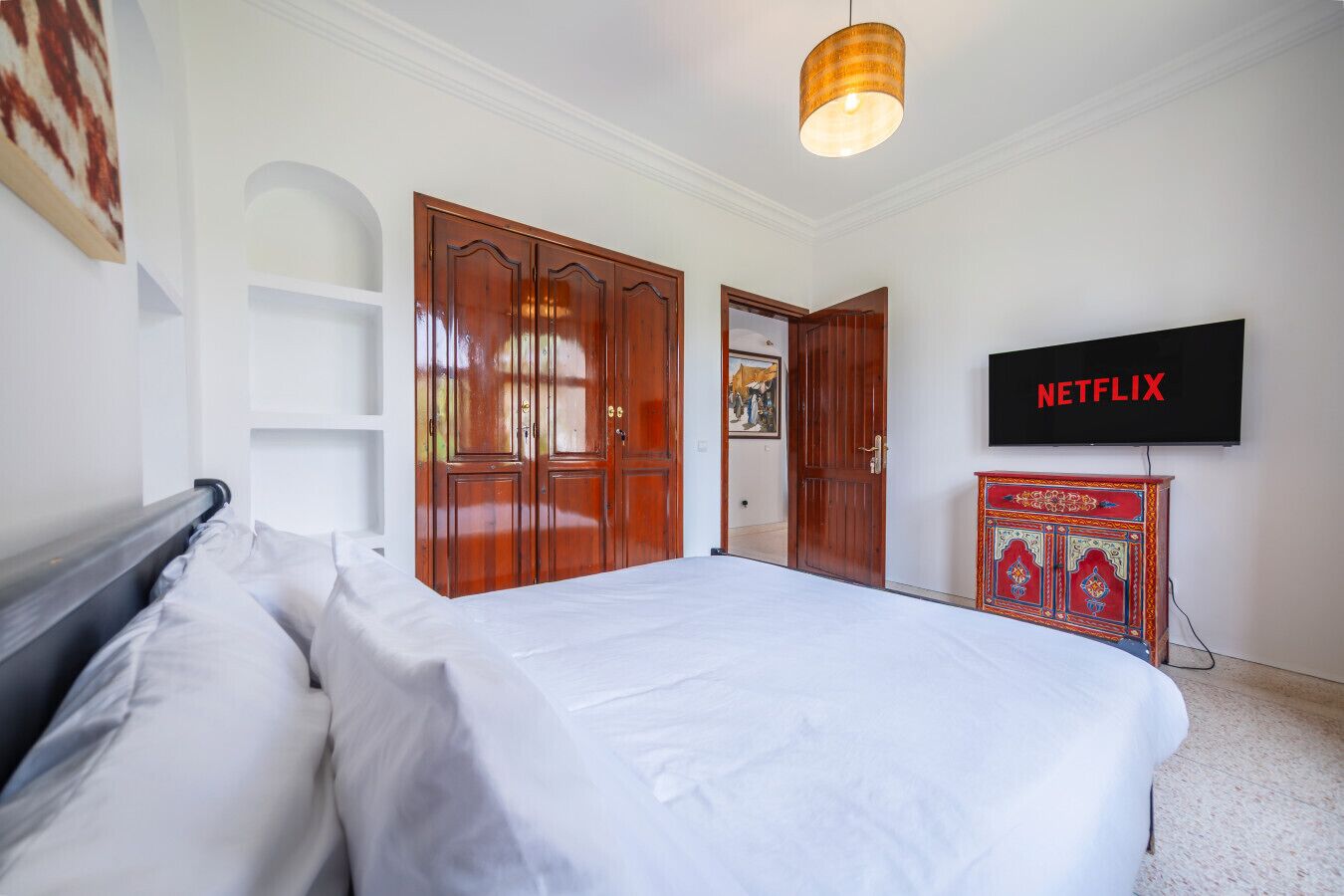 A bedroom with a bed, wooden wardrobe, a TV mounted on the wall, and a shelf unit.