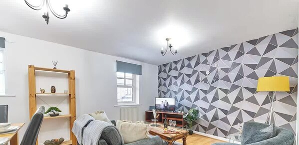 A living room with a sofa, a coffee table, a TV, a wooden shelf, and a geometric-patterned wall. - Modern 2Bed Home - Contractors & Family - Parking (Tyne and Wear)