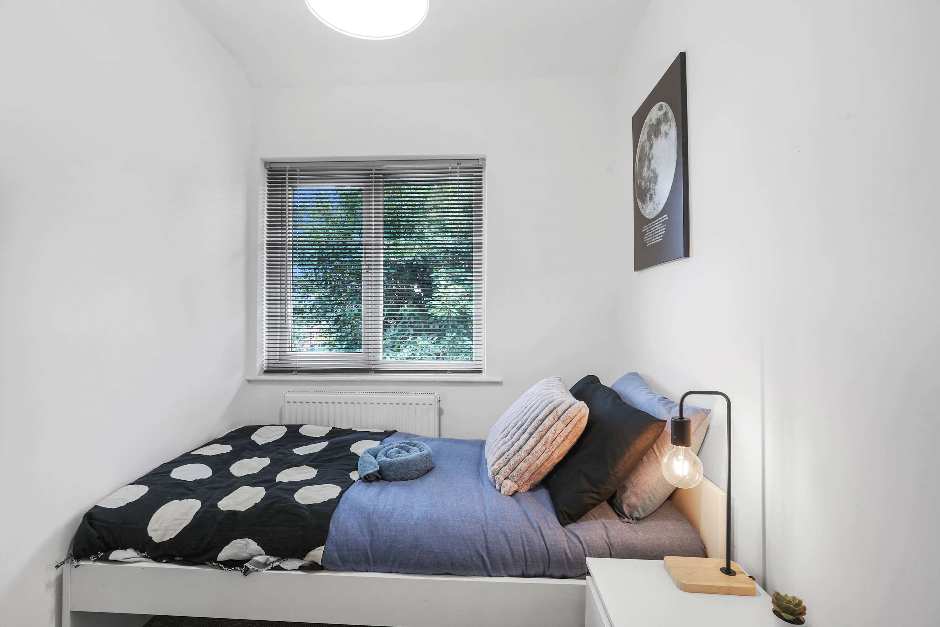 A bedroom with a bed, a nightstand, a lamp, a window with blinds, and a framed picture on the wall.