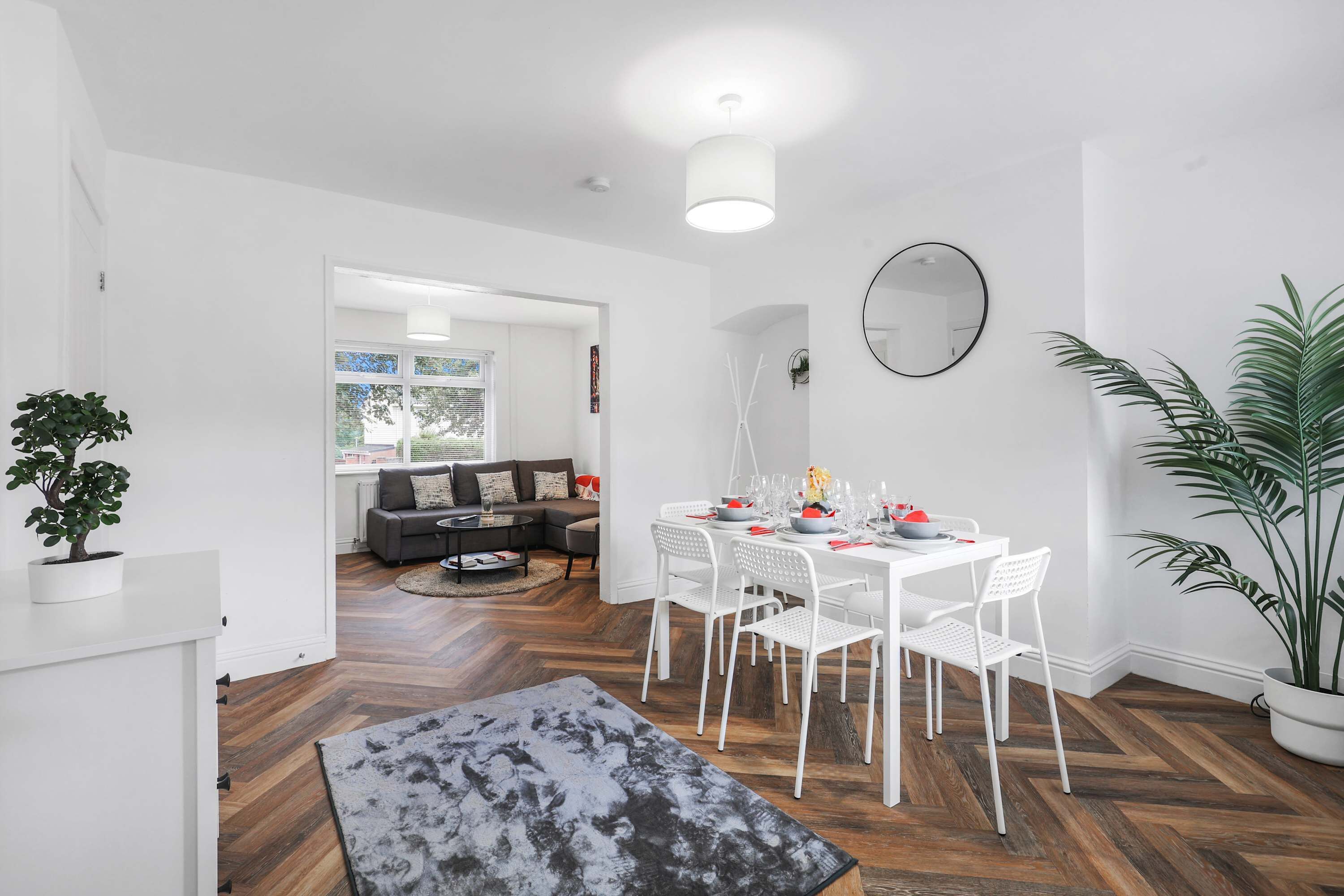 A modern living room with a white dining table set for four, a grey sofa, and a large potted plant.