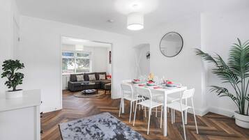 A modern living room with a white dining table set for four, a grey sofa, and a large potted plant.