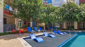 Ein Poolbereich mit blauen Liegestühlen und roten Stühlen, umgeben von Bäumen und Gebäuden.