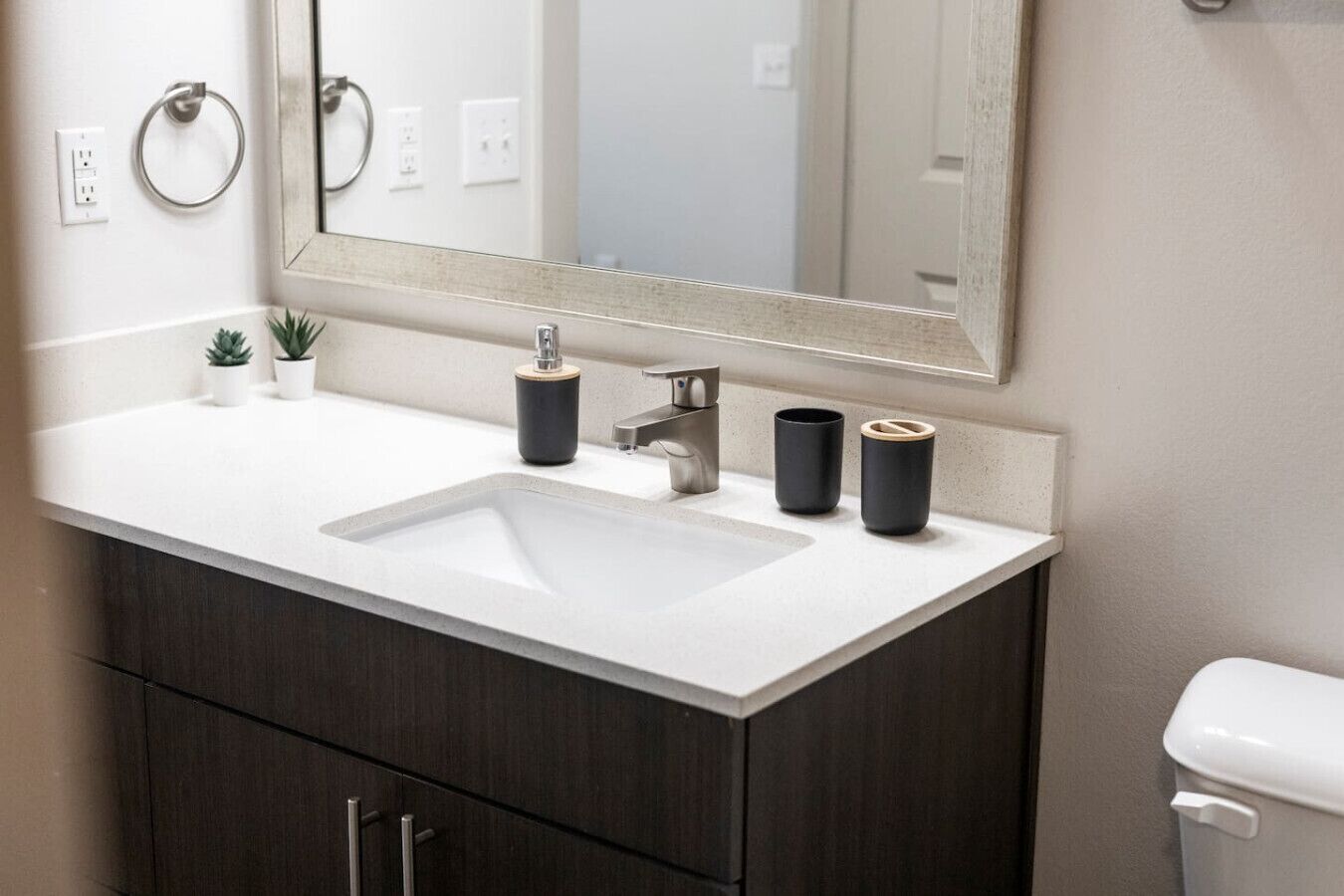A modern bathroom with a white sink, dark wood cabinets, a large mirror, and two black cups on the counter.