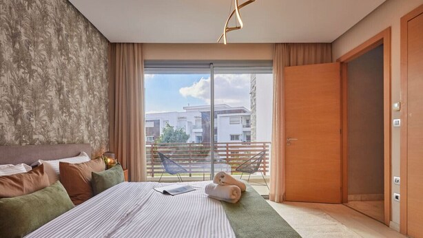 A modern hotel room with a large bed, a view of the city through a sliding glass door, and a balcony with a chair. - Luxury 3-Bedroom Getaway in Bouskoura`s Finest (Bouskoura)