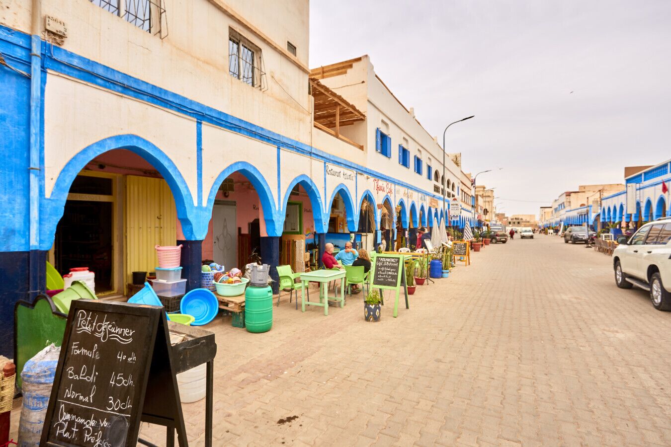 A street with blue-painted buildings, outdoor seating, and a chalkboard sign listing prices.